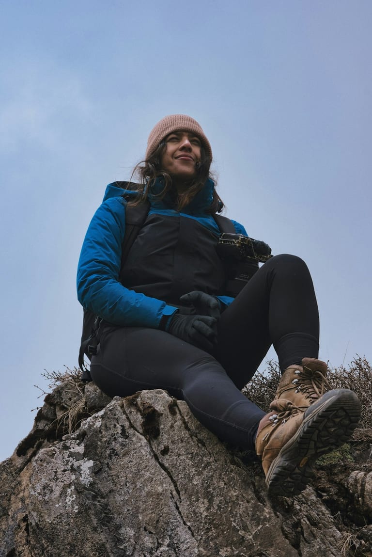 Person sitting on rock outcrop wearing blue jacket and pink beanie, looking upward against clear sky.