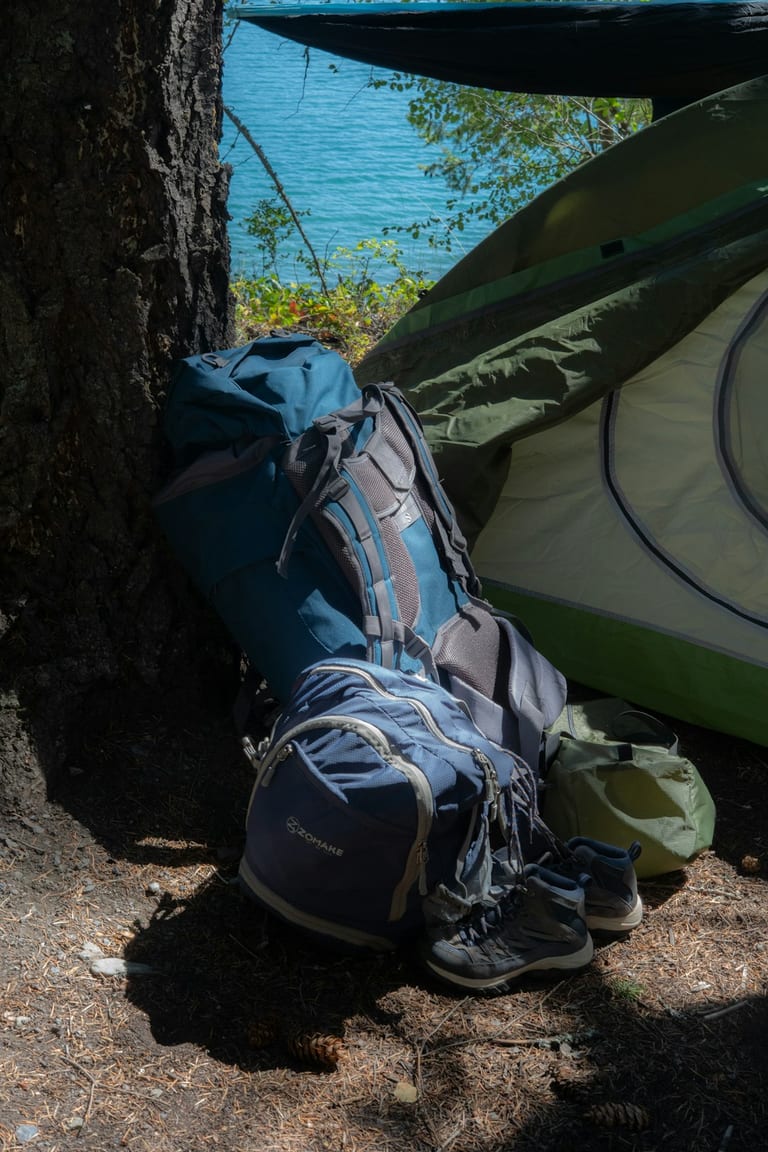 Hiking backpack and shoes rest on ground near a tree, with blue water and green boat visible in background.